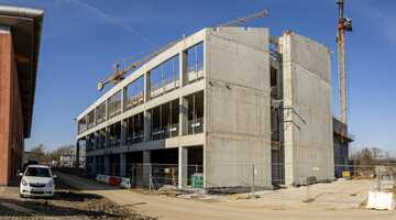 Zdjęcie przedstawiające średni etap budowy nowoczesnego budynku. Na pierwszym planie widoczne jest ogrodzenie budowy, a za nim wielopoziomowy budynek z betonu z rusztowaniami, dźwigiem i pracownikami na rusztowaniach.