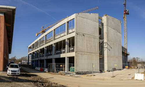 Zdjęcie przedstawiające średni etap budowy nowoczesnego budynku. Na pierwszym planie widoczne jest ogrodzenie budowy, a za nim wielopoziomowy budynek z betonu z rusztowaniami, dźwigiem i pracownikami na rusztowaniach.